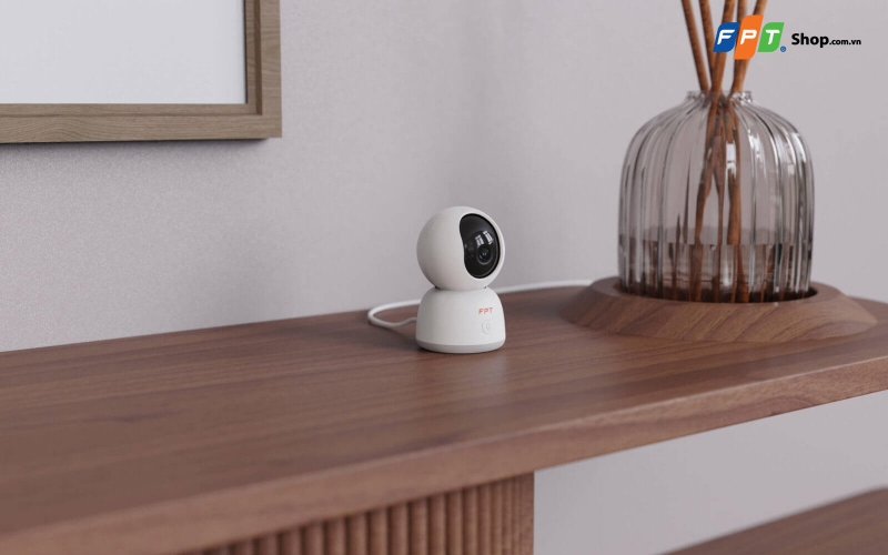 Camera giám sát màu trắng đặt trên kệ gỗ cạnh bình trang trí
