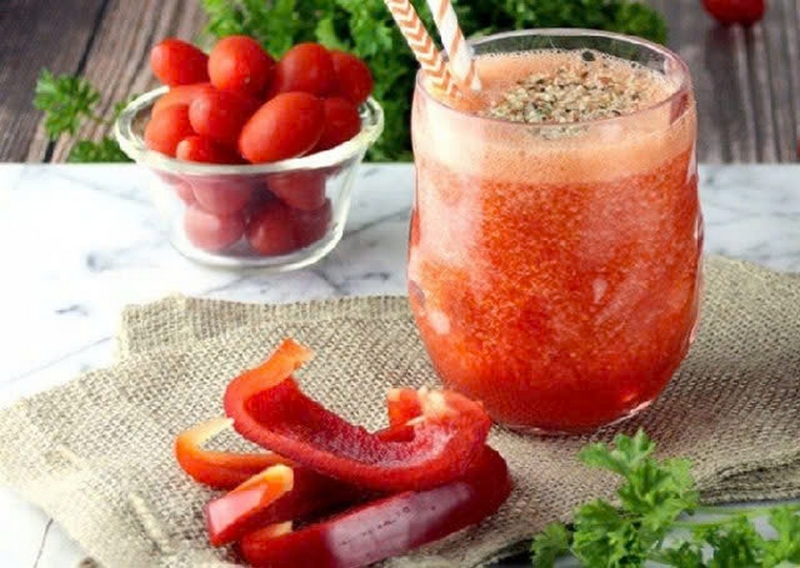 Ly nước ép cà chua và ớt chuông trên bàn, cạnh bát cà chua và lát ớt chuông.