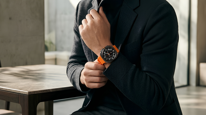 Người đàn ông mặc blazer tối màu đang chỉnh tay áo, để lộ chiếc Galaxy Watch Ultra dây cam trên cổ tay, bên chiếc bàn đá trong không gian tối giản.