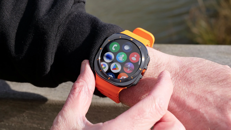 Người đeo Galaxy Watch Ultra dây cam đang dùng ngón tay thao tác trên màn hình hiển thị các ứng dụng ngoài trời.