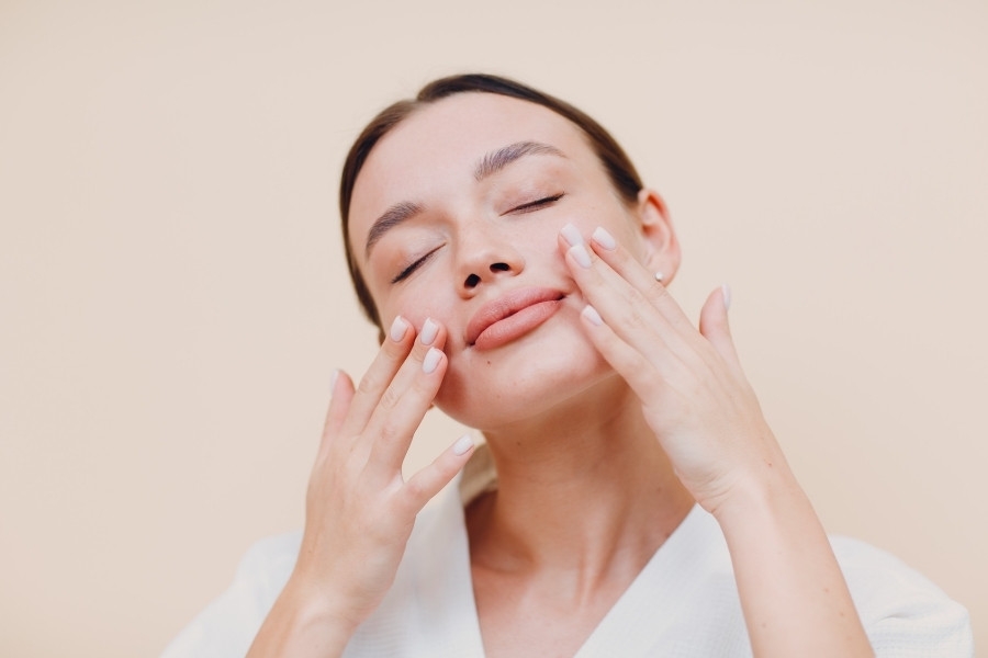 Người phụ nữ thư giãn, hai tay chạm nhẹ lên mặt, mắt nhắm, trên nền màu be nhạt.